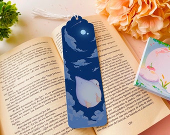May include: A blue bookmark with a white bird flying in a night sky with a crescent moon and stars. The bookmark has a white tassel.