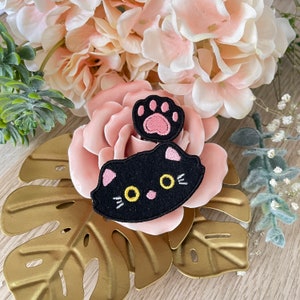 Puede incluir: Una aplicación de gato de fieltro negro con ojos amarillos y nariz rosa. El gato está sobre una flor de fieltro rosa con una huella de pata de fieltro rosa en la parte superior.