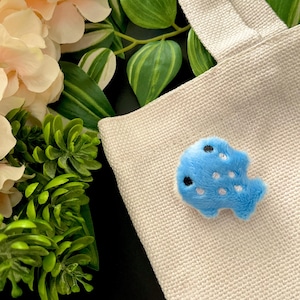 May include: A light blue plush fish applique with white spots and black eyes is attached to a beige canvas tote bag. The fish is made of soft, fuzzy material. The background includes greenery and flowers.