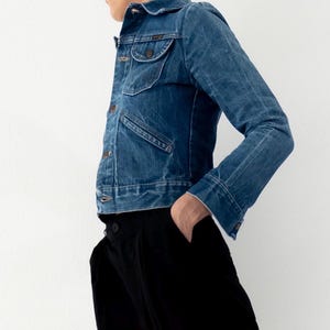 Op de afbeelding: Een spijkerjasje in een medium wassing met twee borstzakken en knoopsluitingen. Het jasje wordt gecombineerd met een zwarte broek. De outfit staat tegen een effen witte achtergrond, wat het contrast tussen denim en de donkere broek benadrukt.