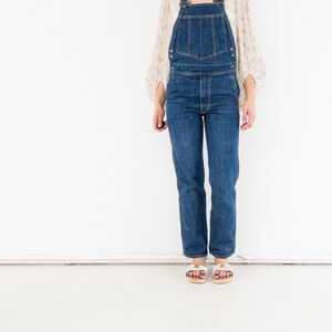 Peut inclure: Une femme portant une salopette en jean bleu avec un chemisier blanc. Elle porte des sandales blanches avec une plateforme en bois.