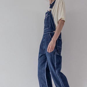 May include: Dark blue denim dungarees featuring a bib pocket, side buttons, and utility pockets. The legs are cuffed at the bottom, revealing a straight-leg cut. A plain cream-coloured short-sleeve t-shirt is worn underneath the denim dungarees.