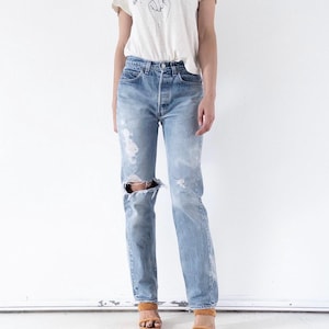 May include: A cream-colored short-sleeve t-shirt with the text "What's Your Sign?" and a graphic of a hand pointing to a crescent moon, paired with light-wash blue denim jeans featuring a distressed knee and tan open-toe heeled sandals.