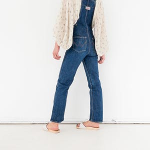 Peut inclure: Une personne portant une salopette en jean bleu avec un chemisier blanc &agrave; motif floral. La salopette a une jambe large et est port&eacute;e avec des sandales blanches.