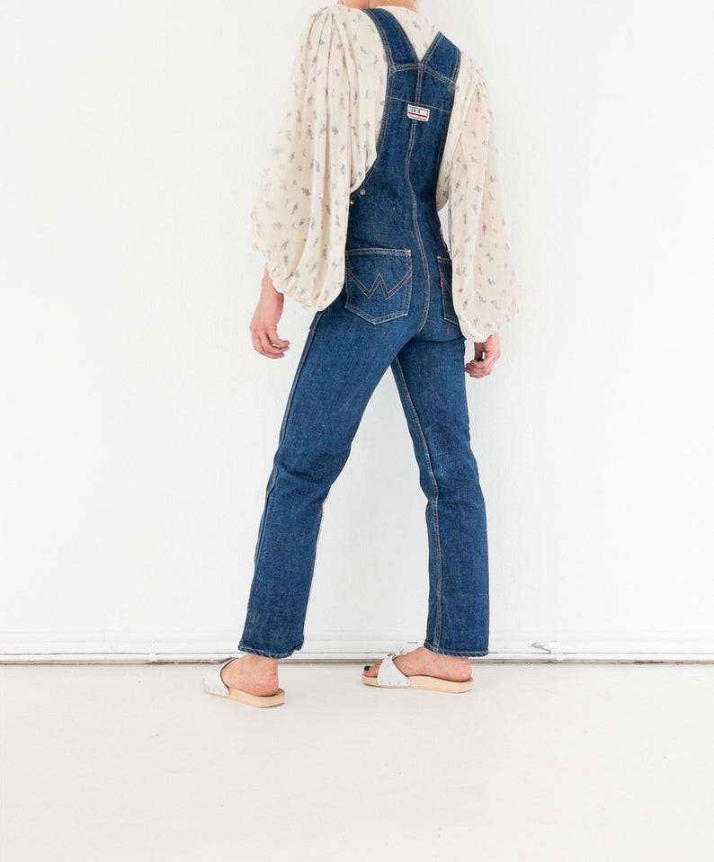 Peut inclure: Une personne portant une salopette en jean bleu avec un chemisier blanc &agrave; motif floral. La salopette a une jambe large et est port&eacute;e avec des sandales blanches.