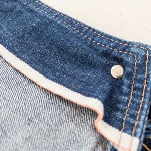 Peut inclure: Gros plan de l'int&eacute;rieur d'une veste en jean bleue, montrant les coutures et un bouton en m&eacute;tal.