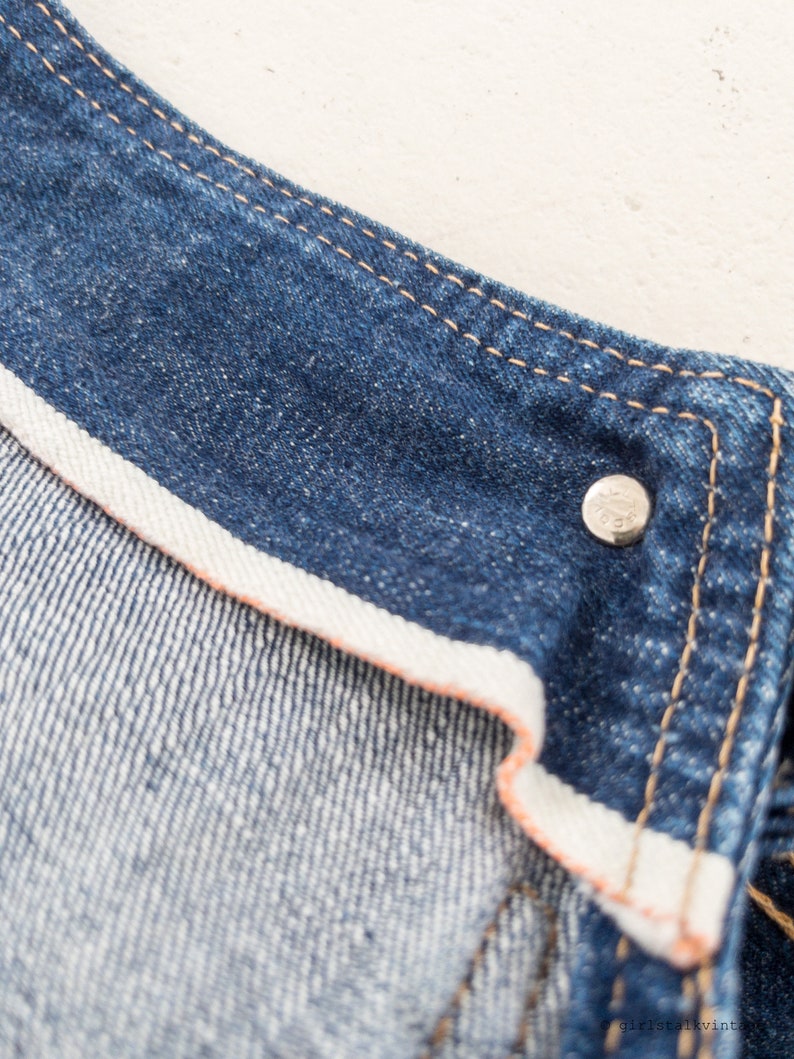 Peut inclure: Gros plan de l'int&eacute;rieur d'une veste en jean bleue, montrant les coutures et un bouton en m&eacute;tal.