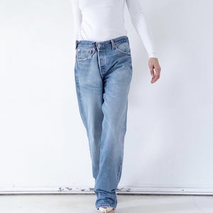 May include: A long-sleeved white crew neck shirt paired with light blue denim jeans. The jeans feature a classic five-pocket design, a button fly closure, and a straight-leg cut with a relaxed fit, showcasing a faded wash and visible stitching.