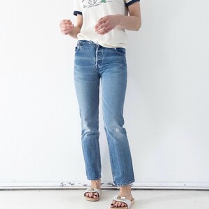 Può includere: Una t-shirt ringer color crema con bordi blu scuro caratterizzata da una grafica di un personaggio dei cartoni animati che scia, abbinata a jeans blu a lavaggio medio con taglio dritto e chiusura a bottoni, e sandali bianchi.