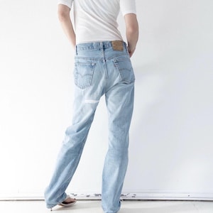 Könnte beinhalten: Eine hellblaue Jeans aus Denim im klassischen Five-Pocket-Design mit einem beigen Lederaufnäher am hinteren Bund und einem kleinen roten Etikett an der rechten Gesäßtasche. Die Jeans hat einen geraden Schnitt und wird mit einem weißen T-Shirt kombiniert.