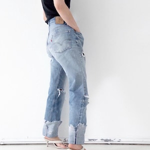Peut inclure: Jean en denim bleu clair avec une coupe taille haute, des détails usés avec des déchirures sur les jambes et de larges revers effilochés aux chevilles. Le jean présente une étiquette en cuir beige à la taille et une petite étiquette rouge.