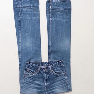Può includere: Jeans in denim blu con un classico design a cinque tasche. I jeans sono stesi piatti, mostrando la parte anteriore e posteriore. Il denim ha un lavaggio sbiadito, con dettagli di usura visibili.