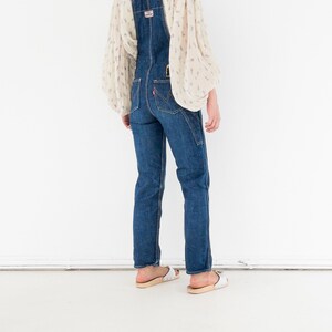 Peut inclure: Une personne portant une salopette en jean bleu avec un chemisier blanc &agrave; motif floral. La salopette a un design classique avec des bretelles r&eacute;glables et une bavette.
