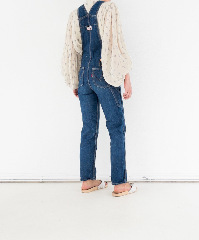 Peut inclure: Une personne portant une salopette en jean bleu avec un chemisier blanc &agrave; motif floral. La salopette a un design classique avec des bretelles r&eacute;glables et une bavette.