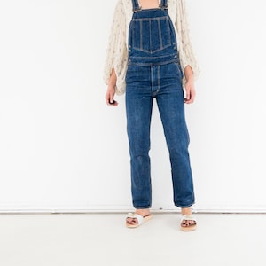 Peut inclure: Une femme portant une salopette en jean bleu avec un chemisier blanc &agrave; fleurs et des sandales blanches.
