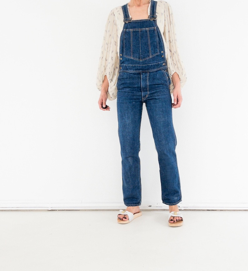 Peut inclure: Une femme portant une salopette en jean bleu avec un chemisier blanc &agrave; fleurs et des sandales blanches.