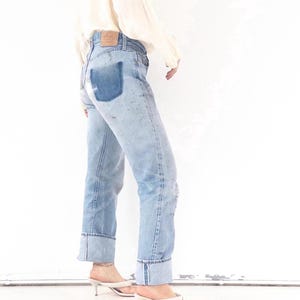 May include: Light blue denim jeans with a patched pocket and rolled cuffs. The jeans have a faded wash and are paired with white heeled sandals. A cream-colored blouse is partially visible.