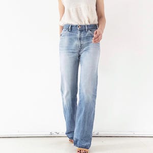 Könnte beinhalten: Ein ärmelloses weißes Oberteil mit hohem Rüschenkragen und strukturiertem, gesmoktem Oberteil, kombiniert mit einer hellblauen Jeans aus Denim. Die Jeans hat einen klassischen Fünf-Taschen-Stil, einen Knopfverschluss und einen geraden Schnitt.