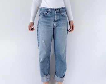 May include: A long-sleeve white crew neck shirt paired with light-wash blue denim jeans featuring a button fly, a high-rise waist, and cuffed hems. The jeans have a relaxed fit with subtle fading and light distressing on the upper thigh area.