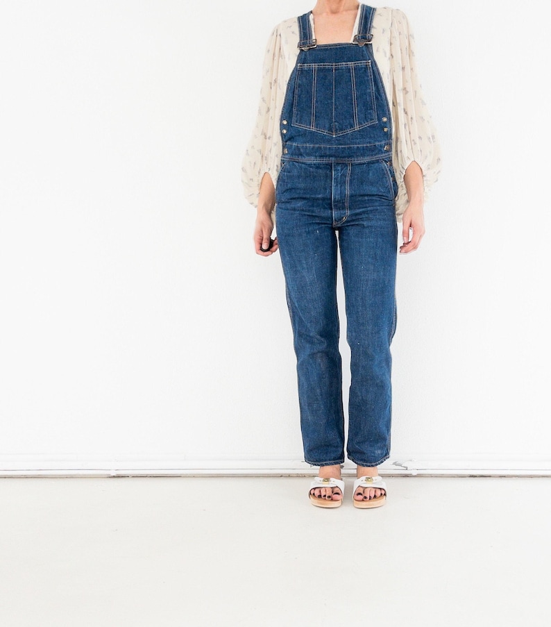 Peut inclure: Une femme portant une salopette en jean bleu avec un chemisier blanc. Elle porte des sandales blanches avec une plateforme en bois.