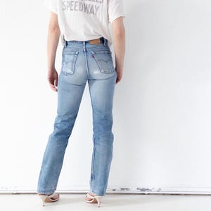 May include: Light blue denim jeans with a faded wash and a classic five-pocket design. The jeans are worn with a white t-shirt that has the text "LOS ANGELES SPEEDWAY" printed on the back. The person is wearing white heeled sandals.