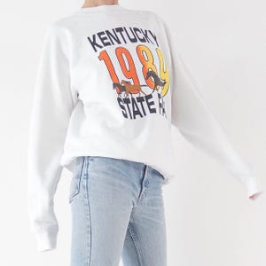 Könnte beinhalten: Weißes Sweatshirt mit langen Ärmeln und dem Text "Kentucky 1984 State Fair" in Schwarz, Orange und Gelb, mit zwei braunen Pferden. Das Sweatshirt wird mit hellblauen Jeans getragen.