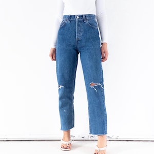 Op de afbeelding: Een persoon draagt een witte top met lange mouwen en een blauwe spijkerbroek met gescheurde knieën en een rafelige zoom. De jeans hebben een hoge taille en een knoopsluiting. De persoon draagt witte sandalen.