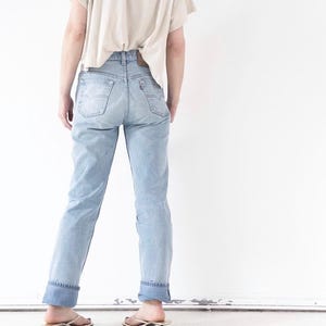 Könnte beinhalten: Eine hellblaue Jeans aus Denim mit hoher Taille, zwei Gesäßtaschen, einem kleinen roten Markenetikett und einem braunen Lederaufnäher am Bund. Die Jeans hat umgeschlagene Säume am Beinabschluss und wird mit einem beigen T-Shirt kombiniert.