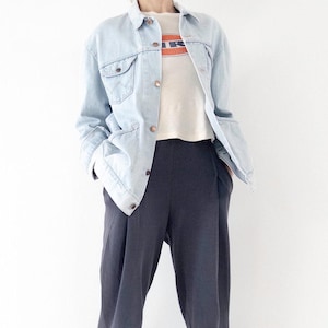 Könnte beinhalten: Hellblaue Jeansjacke mit Knopfverschlüssen und Taschen, kombiniert mit einem cremefarbenen T-Shirt mit orangefarbenem und weißem Aufdruck. Das Outfit wird durch eine dunkelgraue Bundfaltenhose ergänzt, was ein lässiges, aber stilvolles Ensemble ergibt.