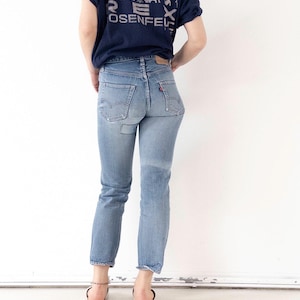 May include: Light blue denim jeans with a red Levi's tag on the back pocket. The jeans have a straight leg fit and are worn with a navy blue t-shirt with a graphic print. The model is wearing sandals.