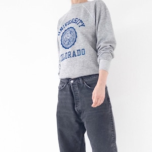 Könnte beinhalten: Ein graumeliertes Langarm-Sweatshirt mit einem blauen Grafikdruck, der University of Colorado und ein kreisförmiges Siegel zeigt. Das Sweatshirt wird mit einer dunkel anthrazitfarbenen Jeans mit Knopfleiste kombiniert.
