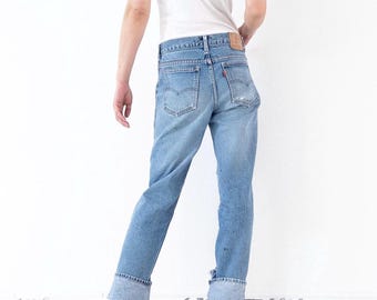 May include: Light blue denim jeans featuring a classic five-pocket design, a tan leather patch on the back waistband, a small red tab on the right back pocket, and wide cuffs at the hem, paired with a plain white short-sleeved t-shirt.