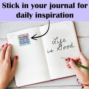 May include: A close-up of an open journal with a colorful sticker that says "We're all just doing the best we can and that is enough." The journal has the words "Life is Good" written in cursive on the right page.