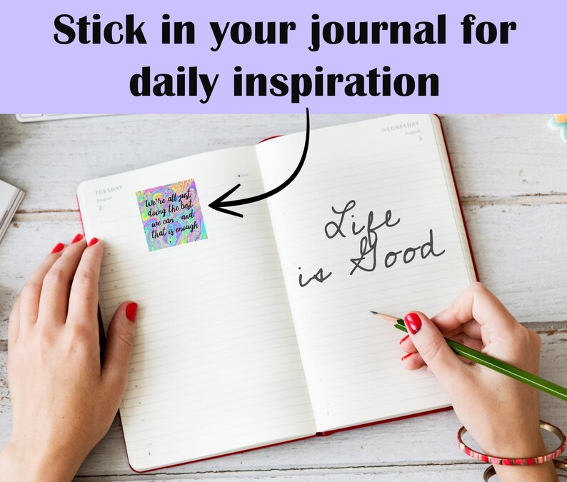 May include: A close-up of an open journal with a colorful sticker that says "We're all just doing the best we can and that is enough." The journal has the words "Life is Good" written in cursive on the right page.