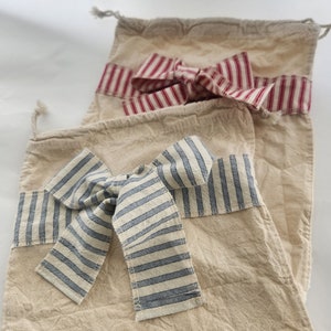 May include: Two rectangular beige fabric drawstring bags. One bag features a horizontal band and a tied bow in red and white stripes. The other bag features a horizontal band and a tied bow in blue and white stripes. Both bags have a textured finish.