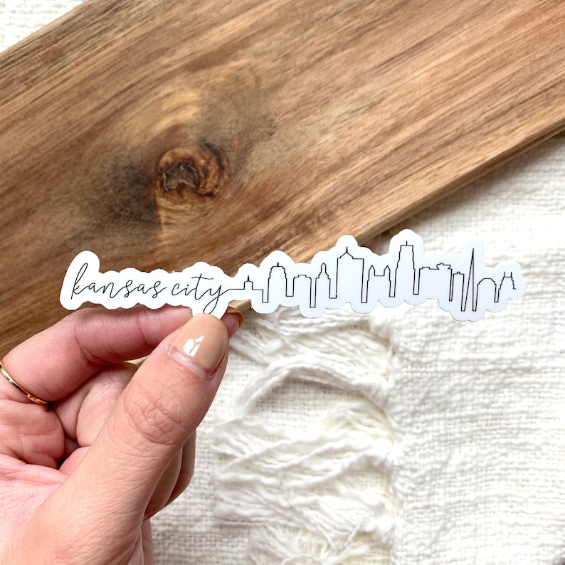May include: A white vinyl sticker featuring a black line art illustration of the Kansas City skyline. The words Kansas City are written in a cursive script font on the left side of the sticker, followed by a continuous outline of city buildings.