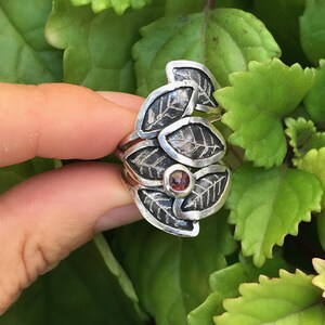 May include: A silver ring with a cluster of leaves and a small red gemstone in the center.