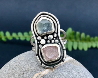 May include: A silver ring with a unique design featuring two gemstones. The top stone is a light blue aquamarine, and the bottom stone is a pink rose quartz. The ring is set in a textured silver band.