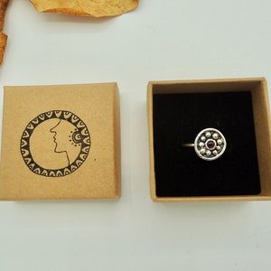 May include: A silver ring with a circular design featuring a small red gemstone surrounded by silver beads. The ring is displayed in a brown box with a black interior.