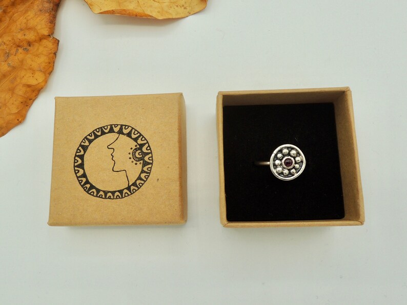 May include: A silver ring with a circular design featuring a small red gemstone surrounded by silver beads. The ring is displayed in a brown box with a black interior.