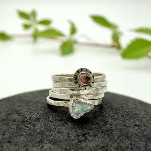 May include: A set of five silver rings with hammered textures. The top ring features a pink gemstone set in a silver bezel with a decorative border. The bottom ring features a light blue gemstone.