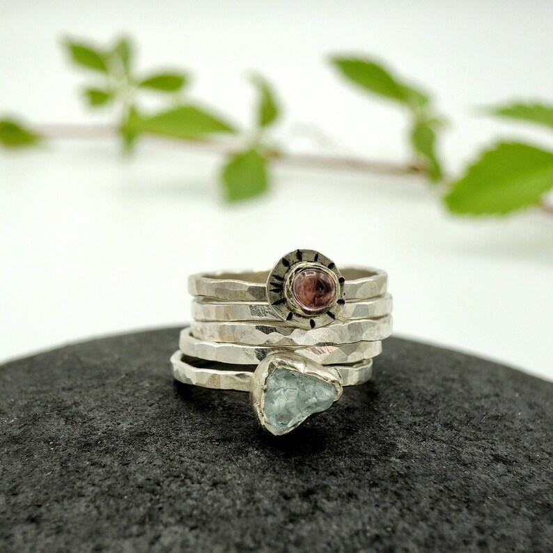 May include: A set of five silver rings with hammered textures. The top ring features a pink gemstone set in a silver bezel with a decorative border. The bottom ring features a light blue gemstone.