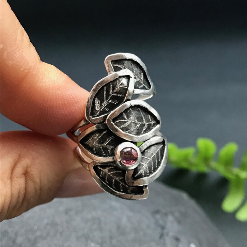May include: A silver ring with a cluster of five leaves, each with a textured surface. A small pink gemstone is set in the center of the ring.