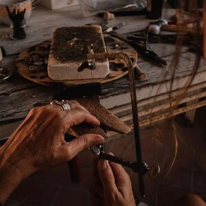 May include: A person's hands are shown using a small hand saw to cut a piece of metal. The metal is resting on a wooden block. The person is wearing several rings on their fingers. The background shows a workbench with various tools and materials.
