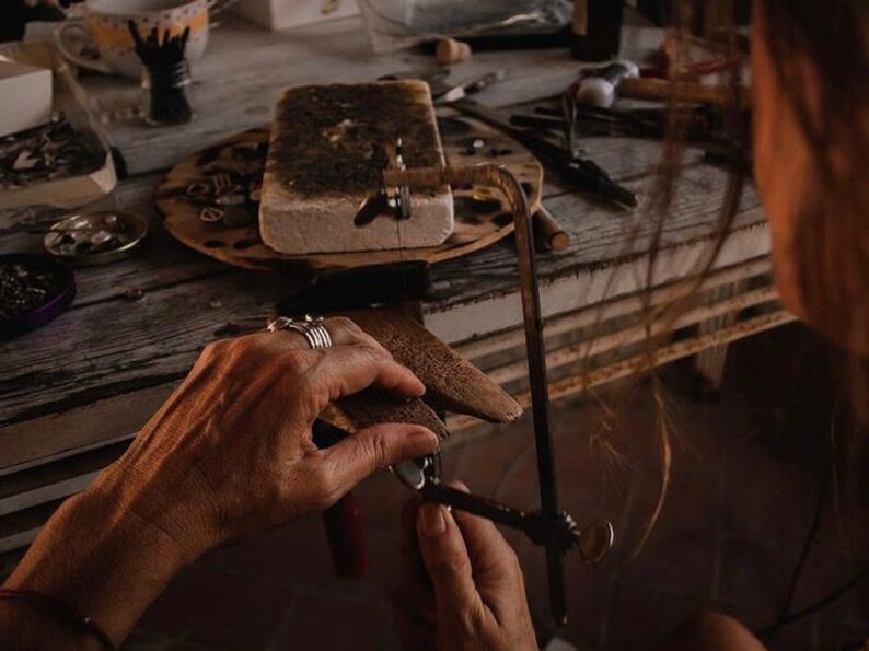 May include: A person's hands are shown using a small hand saw to cut a piece of metal. The metal is resting on a wooden block. The person is wearing several rings on their fingers. The background shows a workbench with various tools and materials.