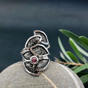 May include: A silver ring with a cluster of five leaves, each with a textured surface, and a pink gemstone in the center.