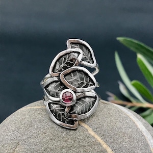 May include: A silver ring with a cluster of five leaves, each with a textured surface, and a pink gemstone in the center.