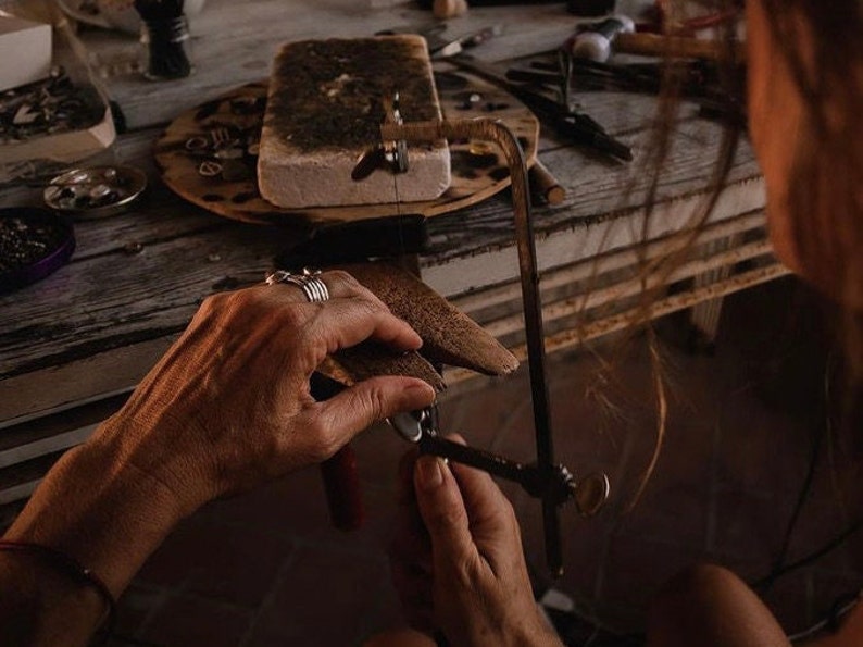 May include: A person uses a hand saw to cut a piece of metal. The metal is clamped in a vise. The person is wearing several rings on their fingers.