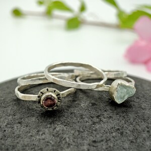 May include: Three silver rings with different stones. The first ring has a small, round, dark red stone. The second ring has a larger, irregular, light blue stone. The third ring is plain.