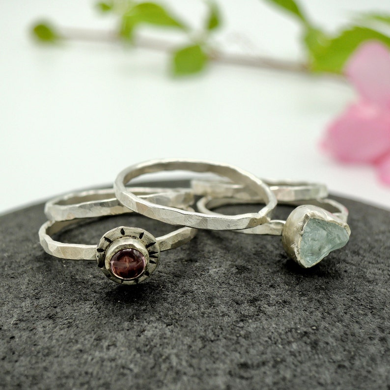 May include: Three silver rings with different stones. The first ring has a small, round, dark red stone. The second ring has a larger, irregular, light blue stone. The third ring is plain.